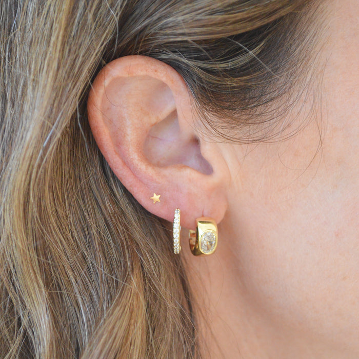 A close-up of a person's ear wearing three gold earrings: a tiny star stud, a huggie hoop with a clear stone, and a textured gold huggie hoop.