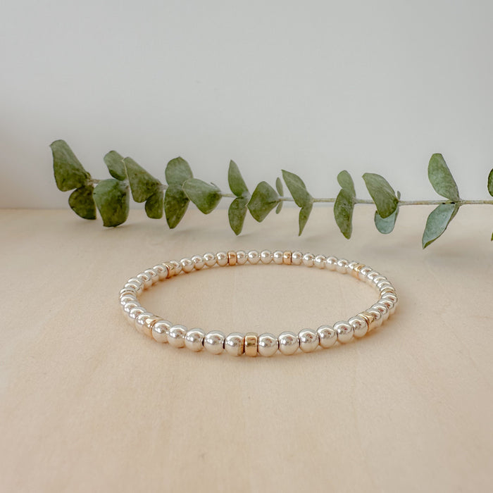 A gold-filled and silver beaded bracelet with a mix of round and rondelle shaped beads, displayed on a wooden surface next to some green leaves for a decorative touch.