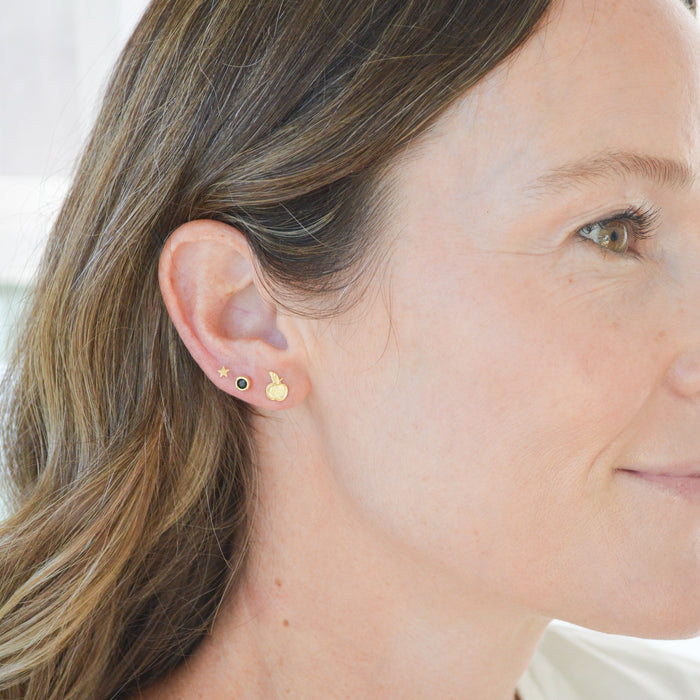 A close-up side profile of a woman wearing two small gold stud earrings, one with a tiny black dot and one with a tiny white dot, on her earlobe.