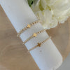 A gold-filled, beaded bracelet with a cross design, displayed on a wooden surface, surrounded by white flowers.