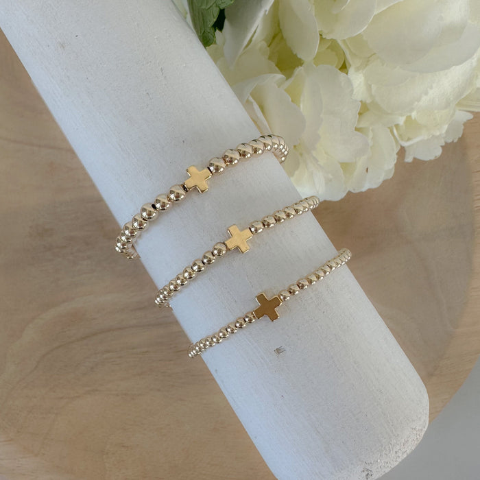 A gold-filled, beaded bracelet with a cross design, displayed on a wooden surface, surrounded by white flowers.
