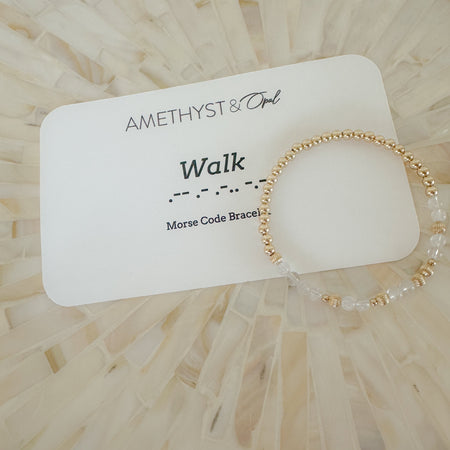 A Morse Code bracelet with the word 'Walk' written above it, featuring 14k gold filled beads and clear gemstone beads on a wooden surface.