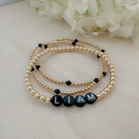 A bracelet with black and white beads, featuring the name 'Liam' in white letters on black beads, displayed on a wooden surface with a flower in the background.