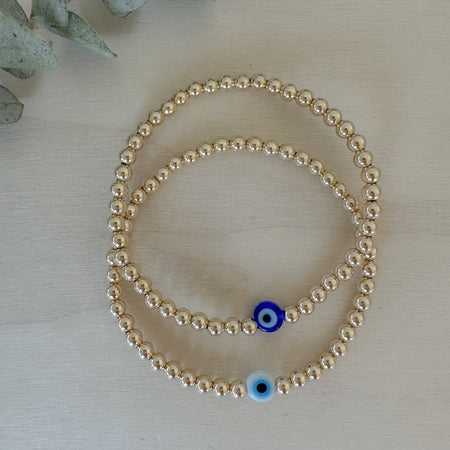 A gold-filled bracelet with a series of uniform beads and a glass bead with a blue evil eye design, strung on a high-quality elastic cord.