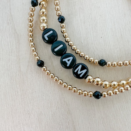 A set of three bracelets, with the top bracelet displaying the name 'LIAM' in white letters on black beads, accompanied by small gold-filled beads.