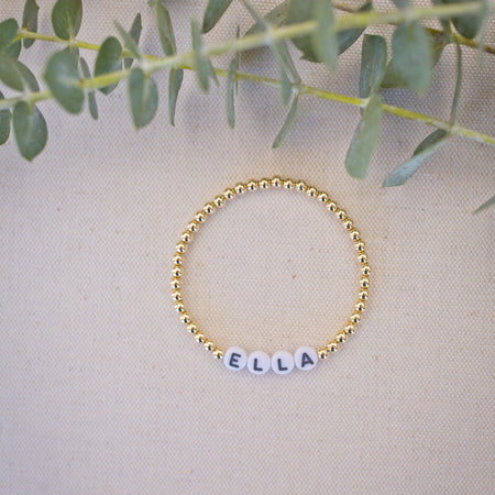 Gold beaded bracelet with name 'Ella' on a beige fabric background