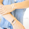 A close-up of a person's hands and arms wearing three gold-tone bracelets: two beaded bracelets with white and black beads, and one bracelet with silver letter beads spelling names.