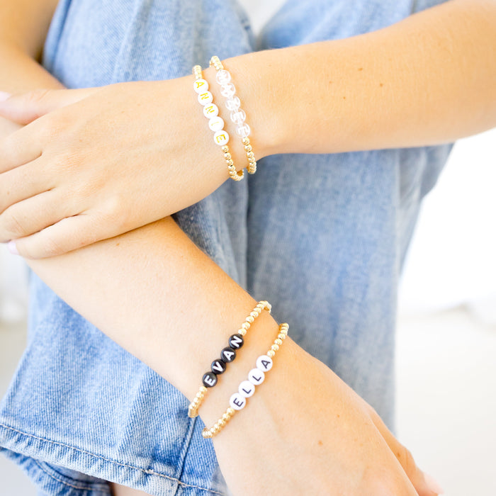 A close-up of a person's hands and arms wearing three gold-tone bracelets: two beaded bracelets with white and black beads, and one bracelet with silver letter beads spelling names.