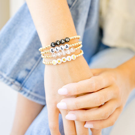 A close-up of a person's wrist and hands wearing three stacked beaded bracelets. The bracelets include black letter beads spelling 'EVA', gold and silver beaded bands, and white letter beads spelling 'ELLA'.