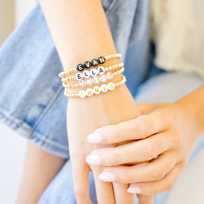 A close-up of a person's wrist and hands wearing three stacked beaded bracelets. The bracelets include black letter beads spelling 'EVA', gold and silver beaded bands, and white letter beads spelling 'ELLA'.