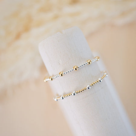 Two gold bracelets on a white cylindrical stand against a beige background