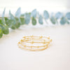 Three delicate gold beaded bracelets with small charm accents, arranged on a light cream surface with blurred green eucalyptus leaves in the background.