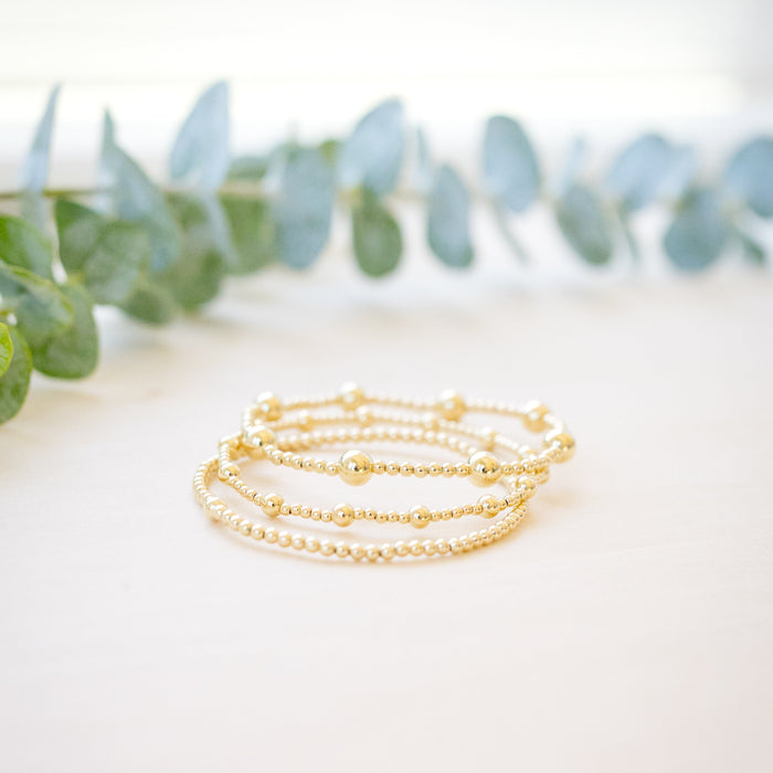 Three delicate gold beaded bracelets with small charm accents, arranged on a light cream surface with blurred green eucalyptus leaves in the background.