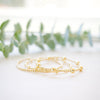 A delicate gold beaded bracelet with a small charm, resting on a white surface with soft-focus green leaves in the background.