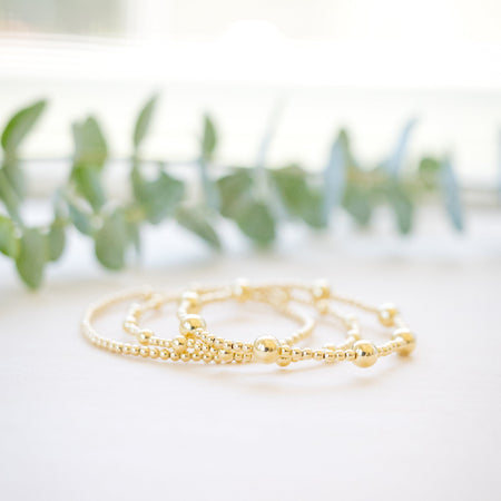 A delicate gold beaded bracelet with a small charm, resting on a white surface with soft-focus green leaves in the background.