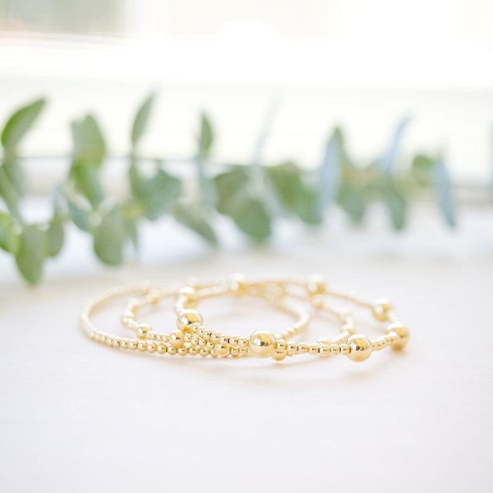 A delicate gold beaded bracelet with a small charm, resting on a white surface with soft-focus green leaves in the background.