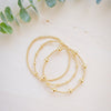 Three delicate gold beaded chain necklaces arranged in a loose spiral on a light beige wooden surface, with soft green eucalyptus leaves framing the top of the scene.