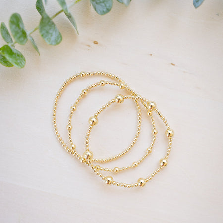 Three delicate gold beaded chain necklaces arranged in a loose spiral on a light beige wooden surface, with soft green eucalyptus leaves framing the top of the scene.