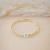 Gold bracelet with a white cord and a small round charm on a beige background