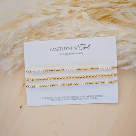 Set of gold and pearl bracelets on a branded card with a textured beige background