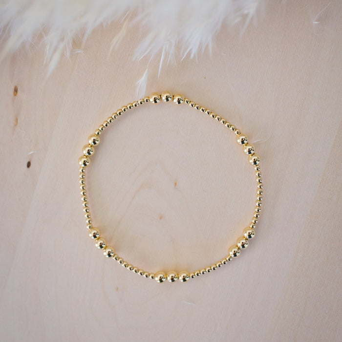 Gold beaded bracelet on a light wooden surface with white feathers