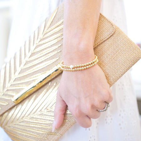 Gold clutch held by a hand with a bracelet, against a white background