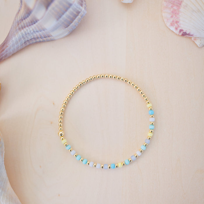 Gold beaded bracelet with blue and yellow beads on a person's wrist against a beach background with shells.