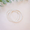 Three delicate silver beaded bracelets arranged in a loose coil on a light beige wooden surface, with soft green eucalyptus leaves in the background.