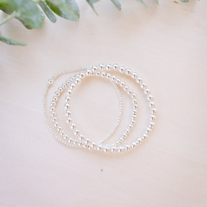 Three delicate silver beaded bracelets arranged in a loose coil on a light beige wooden surface, with soft green eucalyptus leaves in the background.