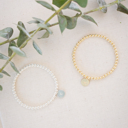 Two bracelets, one gold and one silver, on a light surface with green leaves in the background.