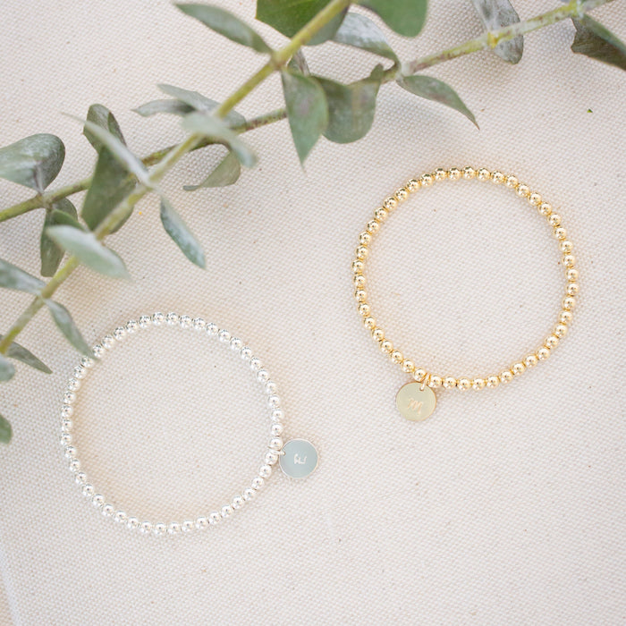 Two bracelets, one gold and one silver, on a light surface with green leaves in the background.