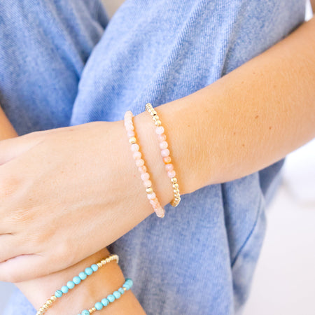 A close-up of a person's hands and legs wearing two beaded stretch bracelets, one in light pink beads with gold spacer beads and the other in light blue/turquoise beads with gold spacer beads, paired with blue jeans.