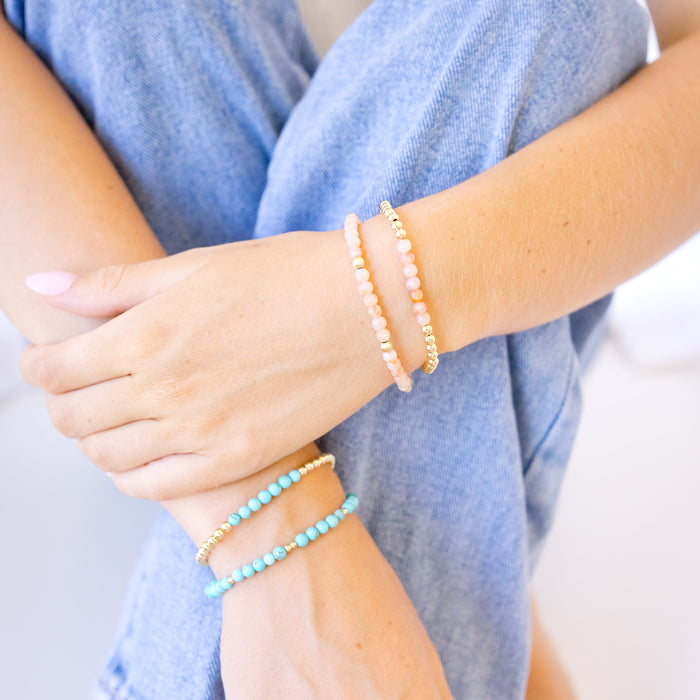 A close-up of a person's hands and legs wearing two beaded stretch bracelets, one in light pink beads with gold spacer beads and the other in light blue/turquoise beads with gold spacer beads, paired with blue jeans.