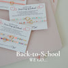 Four small beaded jewelry cards arranged on a pale pink notebook with ribbon bows. The cards read 'First Day of School' with colorful string beads and small heart and bow illustrations.