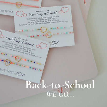 Four small beaded jewelry cards arranged on a pale pink notebook with ribbon bows. The cards read 'First Day of School' with colorful string beads and small heart and bow illustrations.