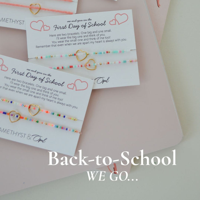 Four small beaded jewelry cards arranged on a pale pink notebook with ribbon bows. The cards read 'First Day of School' with colorful string beads and small heart and bow illustrations.