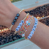 A close-up of a person's wrist wearing multiple beaded bracelets, including one with black letter beads spelling EVAN, one with white letter beads spelling ELLA, and one with white letter beads spelling ELIZABETH, alongside colorful bead strands and gold-tone chains, set against an outdoor patio background with fire pit rocks and warm lights.