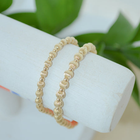 Gold beaded bracelet on a white stand with green leaves in the background