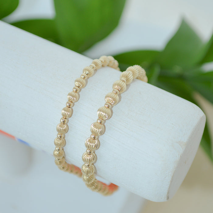 Gold beaded bracelet on a white stand with green leaves in the background
