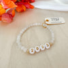 A gold-filled beaded bracelet with white and gold letter/number beads, featuring a moonstone bead, displayed on a table with a colorful flower arrangement in the background.