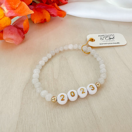 A gold-filled beaded bracelet with white and gold letter/number beads, featuring a moonstone bead, displayed on a table with a colorful flower arrangement in the background.