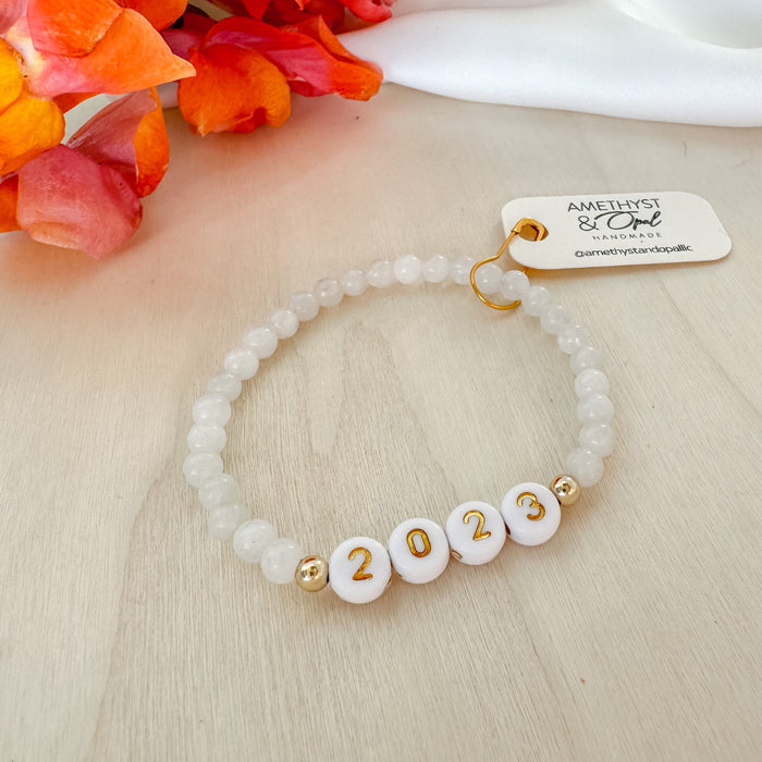A gold-filled beaded bracelet with white and gold letter/number beads, featuring a moonstone bead, displayed on a table with a colorful flower arrangement in the background.