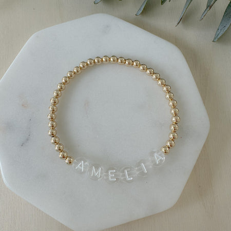 Gold beaded bracelet with 'AMELIA' engraving on a marble surface.