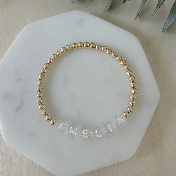 Gold beaded bracelet with 'AMELIA' engraving on a marble surface.
