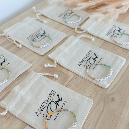 A collection of colorful bracelets displayed on a wooden surface, with each bracelet packaged in a beige branded bag with the name 'Amethyst + Opal'.