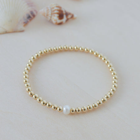 Gold beaded bracelet with a single white pearl on a light background with shells.