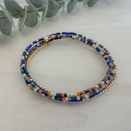 A stack of four bracelets featuring a mix of navy, sage, pumpkin, and brown colors with gold filled beads and delicate seed beadwork on a white wooden surface.