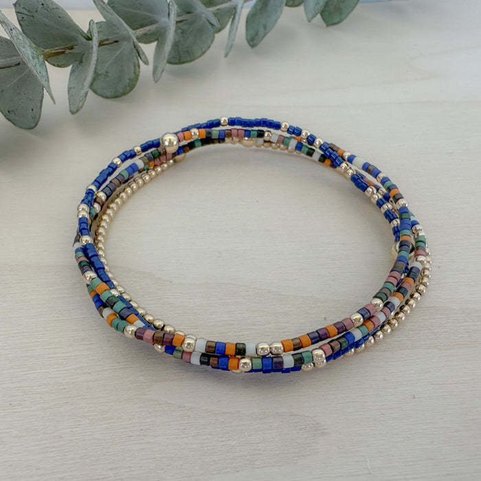 A stack of four bracelets featuring a mix of navy, sage, pumpkin, and brown colors with gold filled beads and delicate seed beadwork on a white wooden surface.