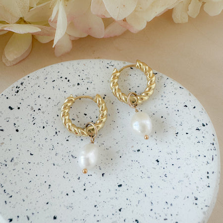 A pair of gold-filled hoop earrings with pearl drops, placed on a white surface with a floral background.