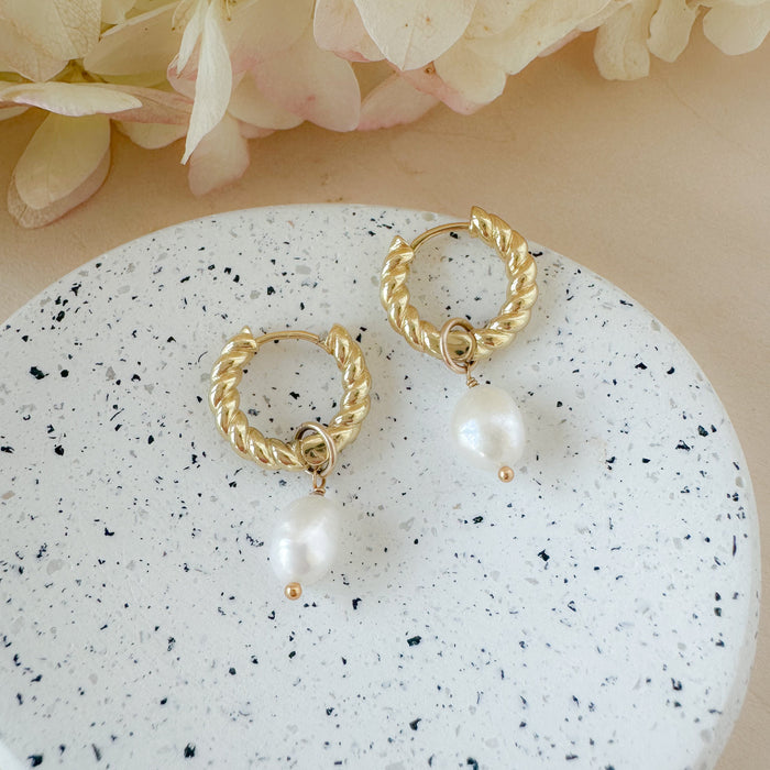 A pair of gold-filled hoop earrings with pearl drops, placed on a white surface with a floral background.